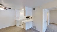 This one-bedroom, one-bathroom unit is designed to maximize functionality within a compact footprint. The kitchen is the central feature of the space, finished in a clean, all-white palette with white cabinetry offering ample storage both above and below the countertops. A built-in sink with a double basin sits beneath upper cabinets, while an electric range with an overhead ventilation hood provides a fully equipped cooking area. A peninsula-style counter extends outward, creating a natural divider between the kitchen and the adjoining living area, and offering additional prep or casual dining space. The flooring transitions from the kitchen's hard surface to neutral wall-to-wall carpeting in the living area, adding warmth underfoot. A textured popcorn ceiling runs throughout, complemented by light beige walls that keep the space feeling bright and open. HVAC vents are positioned strategically on both the walls and floor to ensure comfortable climate control. The overall layout is straightforward and efficient, making practical use of every square foot available on the single floor.