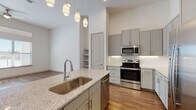 This well-designed single-floor residence showcases a thoughtful open-concept layout that seamlessly connects the kitchen, dining, and living areas. The kitchen serves as the centerpiece of the space, featuring a large island with a granite countertop, undermount stainless steel sink, and a trio of elegant pendant lights overhead. Gray shaker-style cabinetry extends to the notably high ceilings, maximizing storage, while a full suite of stainless steel appliances — including a French door refrigerator, electric range, built-in microwave, and dishwasher — completes the space. A white subway tile backsplash adds a clean, contemporary touch.

Wood-look flooring flows throughout the main living areas, complementing the bright, neutral color palette. The living area benefits from abundant natural light through large windows and a glass door that provides outdoor access, with a ceiling fan adding comfort. Built-in shelving offers additional storage near the kitchen entry.

The bedroom features plush carpeting and a generous window that brings in natural light. The bathroom presents a long vanity with granite countertops, gray cabinetry with brushed nickel hardware, a mirror, and wood-look flooring, along with a separate tub and shower area illuminated by a window.