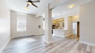 This single-floor unit offers a thoughtfully arranged layout comprising a living room, kitchen, and bathroom. Light gray hardwood-style flooring runs throughout, lending a clean, cohesive look to the entire space. The open-concept living room is bright and airy, featuring neutral warm-beige walls, a ceiling fan with integrated lighting, large windows that allow natural light to pour in, and baseboard heating. A structural column adds a subtle architectural detail that defines the transition between the living area and the kitchen. The kitchen is well-appointed with gray shaker-style cabinetry, white quartz countertops, stainless steel appliances including a refrigerator, microwave, and range, and a peninsula counter that provides additional workspace and casual seating. Pendant and flush-mount lighting illuminate the kitchen with a warm glow. The bathroom is neatly finished with a white vanity featuring a clean countertop and mirror cabinet, wood-style flooring that complements the rest of the unit, and a clean, functional layout. Throughout the space, the consistent use of neutral tones, modern fixtures, and quality finishes creates a bright, welcoming, and practical environment.