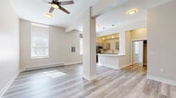 This single-floor unit offers a thoughtfully arranged layout comprising a living room, kitchen, and bathroom. Light gray hardwood-style flooring runs throughout, lending a clean, cohesive look to the entire space. The open-concept living room is bright and airy, featuring neutral warm-beige walls, a ceiling fan with integrated lighting, large windows that allow natural light to pour in, and baseboard heating. A structural column adds a subtle architectural detail that defines the transition between the living area and the kitchen. The kitchen is well-appointed with gray shaker-style cabinetry, white quartz countertops, stainless steel appliances including a refrigerator, microwave, and range, and a peninsula counter that provides additional workspace and casual seating. Pendant and flush-mount lighting illuminate the kitchen with a warm glow. The bathroom is neatly finished with a white vanity featuring a clean countertop and mirror cabinet, wood-style flooring that complements the rest of the unit, and a clean, functional layout. Throughout the space, the consistent use of neutral tones, modern fixtures, and quality finishes creates a bright, welcoming, and practical environment.