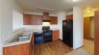 Located at 16102 East Broadway Avenue in Spokane Valley, Washington, this single-floor unit offers a practical and well-organized layout designed for comfortable everyday living. The open living and dining area is finished with neutral wall-to-wall carpeting, warm wood baseboards, and a wall-mounted HVAC unit for individual climate control. Natural light enters through a large window on one side, while a sliding glass door on the opposite end provides direct access to an outdoor area and brings in additional light and exterior views. A pass-through opening connects the living space to the kitchen, creating an open feel between the two areas. The kitchen is equipped with warm-toned wood cabinetry offering generous storage, light-colored countertops, a double stainless steel sink, a black electric range with overhead ventilation hood, a black dishwasher, and a full-size black refrigerator. The kitchen also features durable wood-look flooring. The bathroom includes a full-size tub and shower combo with clean white surround and chrome fixtures. The overall floor plan, visible in the dollhouse view, reveals a straightforward and efficient layout with well-defined living, cooking, and sleeping zones throughout the single level.
