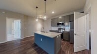 Located at 2460 Walcutt Rd in Columbus, Ohio, this thoughtfully designed single-floor residence offers a clean, contemporary aesthetic throughout. The kitchen serves as a focal point of the home, showcasing gray shaker-style cabinetry, a white subway tile backsplash, granite countertops, and a large center island with an undermount sink. Stainless steel appliances complement the space, while stylish pendant lighting adds a modern touch overhead. Rich hardwood-style flooring flows seamlessly through the kitchen and living areas, creating a cohesive and inviting atmosphere. The living room is bright and open, benefiting from natural light through large windows with views of the surrounding greenery, and features a convenient exterior door. A hallway connects the living spaces to the private areas of the home. Bedrooms are finished with plush carpeting and well-sized windows that allow for ample natural light. Two bathrooms serve the unit: one features a walk-in glass shower with a gray vanity, while the other offers a full soaking tub and shower combination alongside a navy blue vanity with white countertops. Consistent hardwood-style flooring ties the bathrooms together with the rest of the home, reinforcing the clean, modern design throughout.
