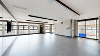 This yoga and fitness studio at Rye House in Baltimore, MD, offers a generously proportioned, open single-floor layout designed to accommodate a variety of workout and wellness activities. The studio's modern aesthetic is defined by clean white walls accented with dark charcoal trim and warm wood detailing along the upper portions of the walls, creating a contemporary yet inviting atmosphere.

A defining feature of the space is the expansive wall of floor-to-ceiling mirrors framed in warm wood trim, providing excellent visibility for form-checking during exercise. Opposite the mirrors, a wide bank of large, multi-pane windows lines the exterior wall, flooding the room with abundant natural light and offering views of the surrounding Baltimore cityscape.

The flooring throughout consists of a durable, textured gray surface well-suited for fitness activities. A large flat-screen display mounted on a tripod stand serves as a focal point for instructor-led or on-demand workout sessions. Additional equipment, including a heavy punching bag, medicine balls on a rack, and stability balls, is available within the space.

Glass-paneled walls and doors connect the studio to adjacent fitness areas, maintaining an open, airy feel. Recessed and linear lighting supplements the natural light, ensuring a well-illuminated environment throughout the day.