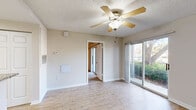 This single-floor unit offers a practical and well-appointed layout across four distinct spaces. The living area features light hardwood-style flooring, neutral walls, a textured ceiling with a ceiling fan, and a sliding glass door that fills the room with natural light while providing views of surrounding greenery. An HVAC vent and wall-mounted thermostat ensure climate control throughout the space.

The kitchen is a functional highlight, featuring white shaker-style cabinetry extending to the ceiling for ample storage, granite-patterned countertops, and a peninsula that doubles as a breakfast bar. Stainless steel appliances—including a range, refrigerator, and range hood—complement the clean white aesthetic, while a recessed ceiling light provides focused illumination.

The bedroom is carpeted for comfort, includes its own ceiling fan, and offers direct outdoor access through a second sliding glass door with views of mature trees. The bathroom adjoins a walk-in closet outfitted with wire shelving systems across multiple levels, providing organized storage. The vanity features a white cabinet base, clean countertop, and a mirrored medicine cabinet. Consistent neutral tones and light finishes throughout create a cohesive, understated ambiance.