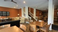This single-floor space makes a strong first impression with its bright, welcoming entryway. The front door is flanked by sidelights that allow natural light to filter in, illuminating patterned flooring and white architectural columns that frame the entry and establish a sense of structure throughout.

The common area beyond is generously sized and open in layout, characterized by warm terracotta-toned accent walls that lend a rich, inviting atmosphere. Recessed lighting and a modern ceiling fan ensure the space remains well-lit and comfortable. A central staircase with a classic white balustrade serves as an architectural focal point, adding visual interest and vertical dimension to the room.

The living area features neutral upholstered seating arranged around a wall-mounted flat-screen television, while a dark countertop bar-style surface provides additional utility. Adjacent dining tables and chairs offer ample seating, making the space well-suited for both relaxed gathering and communal use. Built-in shelving near the staircase adds practical storage and display opportunities. Crown molding and white column accents throughout reinforce a clean, polished aesthetic that ties the entire space together cohesively.