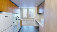 Located at 218 6th Ave in downtown Des Moines, Iowa, this efficiently designed unit offers a well-organized single-floor layout comprising a kitchen, living room, and bathroom. The kitchen is outfitted with warm wood cabinetry — both upper and lower — white appliances including a range, dishwasher, and refrigerator, along with a built-in microwave, a stainless steel sink with a matte black faucet, and clean white countertops. Durable tile-style flooring runs throughout the kitchen area, transitioning to plush carpet in the living space. The living room benefits from two large double-hung windows that frame views of the surrounding downtown cityscape, filling the room with natural light. A built-in counter with overhead cabinetry near the entry provides practical additional workspace or storage. The bathroom is finished in a clean, modern style featuring a trough-style white vanity, a large mirror, a combined tub and shower unit, and wood-look tile flooring. Throughout the unit, warm wood-toned doors and cabinetry provide a consistent design element against the predominantly white walls and ceilings, creating a bright and functional urban living environment.