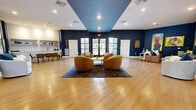 This generously proportioned single-floor space functions as a stylish communal lounge and gathering area, thoughtfully designed with a bold, contemporary aesthetic. The interior is defined by deep navy blue walls that extend across most surfaces, creating a dramatic contrast against the light hardwood flooring that runs throughout the entire space. A central recessed ceiling section, painted in a coordinating teal-blue tone, adds architectural depth and visually anchors the main lounge area below.

The lounge zone is furnished with a curated arrangement of rounded, cognac-toned armchairs surrounding a low white coffee table, all set atop a richly patterned area rug. Multiple white sofas and accent seating pieces are positioned throughout the space, offering varied social configurations. The walls serve as a gallery, displaying a rotating collection of vibrant, large-scale artworks that inject color and cultural character into the room.

Along one side, an open kitchenette and bar area features open wood shelving, a sink, and a long communal table with leather counter-height chairs — ideal for casual collaboration or refreshment. Large black-framed windows and glass double doors allow abundant natural light to pour in, offering exterior views and maintaining an open, airy atmosphere throughout.