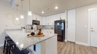 This single-floor residence offers a cohesive, modern design characterized by neutral tones, wood-look plank flooring, and granite countertops carried consistently throughout the space. The open-concept kitchen serves as a central highlight, featuring crisp white shaker cabinetry with matte black hardware, granite countertops, and black appliances including a refrigerator, range, and over-the-range microwave. A large peninsula with seating for three provides both additional workspace and a casual dining area, illuminated by a row of pendant lights overhead.

The living area adjoins the kitchen seamlessly, offering a bright and welcoming atmosphere enhanced by natural light filtering through sheer curtains. Neutral wall tones and clean lines give the space a relaxed yet polished feel.

The two bedrooms feature plush carpeting and generous closet storage. Both are well-proportioned and benefit from natural light through large windows. The two bathrooms mirror the kitchen's aesthetic with white vanity cabinetry, granite countertops, and wood-look flooring. One bathroom offers a walk-in shower, while the other includes a shower-tub combination.

A dedicated laundry room with side-by-side washer and dryer units and additional cabinetry adds notable everyday convenience to this well-appointed space.