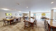 Located at 140 Spencer St in Manchester, CT, this single-floor community space seamlessly blends a dining room and living room into one generously sized, open-plan area. The dining section is furnished with multiple sturdy wood tables and cushioned chairs arranged to accommodate group gatherings, all set atop a patterned, multi-tone carpet that adds warmth and texture to the room. Structural columns with decorative stone bases provide architectural character while defining the layout without enclosing the space.

Natural light flows in abundantly through large windows and a set of French doors, which open directly to an outdoor patio area with seating — offering a pleasant indoor-outdoor connection. The neutral, warm beige walls are accented with framed artwork and wall décor, contributing to a welcoming and calm atmosphere.

The living area transitions naturally from the dining space and features plush sofas and oversized armchairs arranged for comfortable conversation. Decorative wall pieces, including a bird motif and a framed military flag display, add meaningful character to the lounge area. Recessed and flush-mount ceiling lights provide consistent, even illumination throughout. Overall, the space presents a functional yet inviting communal environment well-suited for dining, relaxing, and socializing.