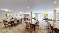 Located at 140 Spencer St in Manchester, CT, this single-floor community space seamlessly blends a dining room and living room into one generously sized, open-plan area. The dining section is furnished with multiple sturdy wood tables and cushioned chairs arranged to accommodate group gatherings, all set atop a patterned, multi-tone carpet that adds warmth and texture to the room. Structural columns with decorative stone bases provide architectural character while defining the layout without enclosing the space.

Natural light flows in abundantly through large windows and a set of French doors, which open directly to an outdoor patio area with seating — offering a pleasant indoor-outdoor connection. The neutral, warm beige walls are accented with framed artwork and wall décor, contributing to a welcoming and calm atmosphere.

The living area transitions naturally from the dining space and features plush sofas and oversized armchairs arranged for comfortable conversation. Decorative wall pieces, including a bird motif and a framed military flag display, add meaningful character to the lounge area. Recessed and flush-mount ceiling lights provide consistent, even illumination throughout. Overall, the space presents a functional yet inviting communal environment well-suited for dining, relaxing, and socializing.