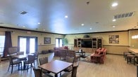This single-floor communal space is designed to serve as a comfortable gathering area, combining a lounge and casual dining zone within one open layout. The room is generously sized, with recessed ceiling lighting illuminating the entire area evenly and crown molding adding a refined architectural detail along the upper perimeter.

The lounge portion of the room is anchored by a large wooden entertainment center housing a flat-screen television, flanked by glass-door cabinetry offering ample display and storage space. Plush, upholstered seating in warm red and orange tones is arranged around a central coffee table atop a decorative area rug, creating an inviting conversational setting. A coordinating sofa and accent chair complete the arrangement nearby.

Toward the front of the room, a dining area features dark wood tables paired with cushioned chairs, suitable for casual meals or social gatherings. Natural light filters in through glass-paneled doors along the wall, enhancing the room's warm, welcoming ambiance. Potted greenery and framed wall art add finishing touches throughout, contributing to the space's comfortable, homey character.