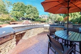 This single-level outdoor patio space is thoughtfully designed to offer a comfortable and functional al fresco environment. The flooring throughout features rich, stamped concrete with a warm brick-toned pattern that adds both texture and visual interest underfoot.

A key architectural element is the curved stacked stone wall that defines the perimeter of the patio, providing both structure and aesthetic charm. Integrated into this wall is a built-in stainless steel grilling station, making it an ideal setting for outdoor entertaining and cooking. Decorative stone pillars topped with ornamental planters and blooming flowers add a refined, welcoming touch to the entryway area.

The dining area is furnished with a round slatted table and high-top chairs, shaded by a large rust-colored market umbrella. An adjacent built-in bench seating area, accented with colorful throw pillows, offers a relaxed lounge space nestled against the brick exterior wall of the building.

Access to the interior is provided through classic French doors fitted with plantation shutters, adding a traditional design detail to the exterior façade. The patio opens up to expansive views of mature trees, manicured landscaping, and neighboring architecture, filling the space with natural light and a strong sense of connection to the outdoors.