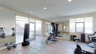 This single-floor fitness room offers a clean, functional environment designed to support a variety of workout routines. The space is finished with large neutral-toned floor tiles complemented by rubber exercise mats, providing a safe and comfortable surface for both equipment-based and floor exercises. A full-wall mirror runs along one side of the room, enhancing the sense of openness while allowing users to monitor their form during workouts. The room is equipped with a multi-station cable and weight machine, an adjustable workout bench, a freestanding punching bag, and yoga or exercise mats, catering to strength training, functional fitness, and flexibility work alike. Recessed ceiling lighting ensures even illumination throughout the space, while multiple large windows and a glass-panel door flood the room with abundant natural light and offer views of the surrounding exterior. The bright, airy ambiance created by the generous fenestration makes the space feel welcoming and energizing. The overall design is practical and straightforward, prioritizing utility and comfort for regular fitness use.