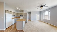 This thoughtfully laid-out 1-bedroom, 1-bathroom residence offers a comfortable and functional living experience across a single floor. The open-concept design seamlessly connects the kitchen and living room, creating a spacious, airy feel throughout the main living area. The kitchen is well-appointed with gray cabinetry, warm-toned countertops, a center island with additional counter space, a white electric range, dishwasher, and durable hardwood-style flooring. A tall pantry cabinet provides generous storage. The living room is expansive and bright, featuring plush carpeting, a ceiling fan with light fixture, and multiple large windows that flood the space with natural light and offer views of the surrounding neighborhood. A glass-panel entry door adds to the welcoming atmosphere. The bedroom is a comfortable retreat with soft carpeting, a ceiling fan, and a window for natural light. The full bathroom features a gray vanity with a warm-toned countertop, a large mirror, and hardwood-style flooring, maintaining the cohesive design aesthetic found throughout the home. Neutral gray walls and clean white trim provide a modern, move-in-ready backdrop throughout the entire residence.