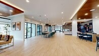 Located at 2100 Marquis Pkwy in Williamsburg, Virginia, this single-floor community space offers a generously proportioned, open-concept layout designed for both functionality and modern aesthetic appeal. The entrance greets visitors with light wood-tone flooring, a warm slatted wood ceiling accent, ambient LED lighting, and a reception desk set against a textured feature wall with contemporary branding — establishing an immediately polished first impression. The expansive main area flows seamlessly from the entrance into a combined living and dining space. The dining zone features a long communal table with teal upholstered chairs beneath dramatic pendant lights, while the living area showcases a rich navy built-in shelving wall, warm wood-paneled ceiling, a stone accent wall, and a large sectional sofa — creating a cozy yet sophisticated lounge. Abundant natural light pours in through large black-framed windows, offering outdoor views of the surrounding neighborhood. The kitchen area includes sleek white cabinetry with gold hardware, marble-style countertops, and a black matte sink. Private glass-walled meeting rooms with black framing provide enclosed spaces for focused work or meetings. Throughout, light hardwood-style flooring, recessed lighting, and a neutral white palette unify the space with a clean, contemporary character.