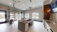 This contemporary single-floor amenity space is designed for socializing and gathering, offering a bright, open-plan layout finished with large-format light tile flooring throughout. At the heart of the room sits a substantial granite-topped island with built-in white cabinetry and bar seating, making it an ideal hub for entertaining. Adjacent to the island, a dedicated kitchenette features white lower cabinets, a stainless steel under-counter refrigerator, a sink, and a wood-paneled accent wall mounting a large flat-screen television flanked by decorative sconce lighting, adding warmth and texture to the modern palette. The space benefits from generous ceiling height, recessed lighting, and a ceiling fan that enhances the airy ambiance. A full-panel glass roll-up garage door serves as a dramatic architectural feature, seamlessly connecting the interior to a covered outdoor patio furnished with round bistro tables and woven rattan chairs. Additional fixed windows along the perimeter flood the interior with natural light and offer views of the surrounding landscaped grounds. The overall design balances a clean, neutral color scheme with warm wood accents, creating a polished yet inviting environment well-suited for community events and casual gatherings.
