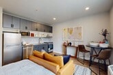 Located at 1600 N 11th St in Philadelphia, PA, this thoughtfully designed one-bedroom, one-bathroom unit offers a smart, open-concept layout that maximizes every square foot. The main living area seamlessly integrates the bedroom, living, dining, and kitchen spaces into one cohesive room, finished with warm wood-look plank flooring and neutral-toned walls that create a bright, welcoming atmosphere. Recessed lighting throughout ensures consistent illumination, while a large window allows natural light to fill the space and provides views of the surrounding neighborhood. The kitchen is efficiently outfitted with slate-gray upper and lower cabinetry, open shelving, a stainless steel range, a full-size refrigerator, a built-in microwave, and a single-basin sink — providing all essential amenities in a compact footprint. A cozy dining nook with a bistro-style table and chairs sits adjacent to the kitchen, making the most of the open layout. A short hallway leads to the private bathroom, which features light tile flooring, a floating white vanity with modern black hardware, and a well-lit mirror. The clean, contemporary design throughout reflects a commitment to functional, modern urban living.