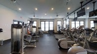 This fitness center offers a comprehensive workout environment thoughtfully organized across a single open floor. The space is designed with functionality and comfort in mind, featuring a neutral color palette of warm beige walls and a dark rubberized flooring that provides both durability and safety during exercise. Recessed lighting paired with ceiling fans ensures the room remains well-lit and properly ventilated throughout the day.

Natural light streams in through multiple large windows along the far wall, creating an inviting atmosphere that enhances the overall workout experience. Along one side of the room, a row of Precor cardio machines — including treadmills and ellipticals — are neatly arranged, each equipped with individual monitors. The opposite side houses a variety of strength training machines and cable systems, offering a full range of resistance exercises. A dedicated free weights area features a tiered dumbbell rack alongside a flat bench, catering to those seeking versatile strength training options. Mirrored wall sections help visually expand the space while allowing users to monitor their form. The layout promotes efficient circulation, making this a practical and well-rounded facility.