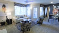 This single-floor space presents a thoughtfully designed living area that balances comfort and style. The room is anchored by a sleek white sofa accented with patterned throw pillows, complemented by a pair of curved accent chairs upholstered in a bold floral motif. A round glass-topped coffee table serves as the centerpiece, resting atop a textured area rug that defines the seating arrangement. Natural light pours in generously through multiple large windows dressed with coordinating Roman shades in a dark circular pattern, creating a cohesive aesthetic throughout the space. A glass-paneled door provides direct access to an outdoor area, where patio furniture is visible, further extending the sense of openness. The ceiling features a ceiling fan, contributing to the room's practical functionality. Neutral carpeted flooring runs throughout, providing a soft, understated base that allows the furnishings to take center stage. The overall ambiance is contemporary and inviting, with a carefully curated color palette of grays, whites, and blacks lending the space a polished, unified appearance.