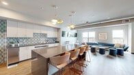 Located at 2050 Durant Ave in Berkeley, CA, this thoughtfully designed communal space combines a fully equipped kitchen and an inviting living area within a single open-plan floor. The kitchen is appointed with sleek white cabinetry, dark countertops, and a striking teal geometric-patterned tile backsplash that adds a vibrant design accent. A large central island with a smooth taupe surface and a row of modern upholstered bar stools serves as both a functional workspace and a social gathering point. Distinctive geometric pendant lights hang overhead, enhancing the contemporary aesthetic. Appliances include a retro-style black refrigerator and stainless steel units, blending style with practicality. The living area flows naturally from the kitchen and features a curved, deep-blue sectional sofa complemented by coordinating blue ottomans and a organic-shaped wood coffee table. A wall-mounted flat-screen TV and an additional dual-screen media setup provide entertainment options, while large windows flood the space with natural light and offer views of the surrounding Berkeley cityscape. Warm wood-tone flooring runs throughout, tying both zones together cohesively. The adjacent hallway features open shelving, greenery, and access to additional rooms, completing this versatile and well-appointed communal environment.