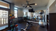 This fitness center is designed to support a comprehensive workout experience within a compact, well-organized layout. The space is divided into two interconnected zones separated by elegant French doors with frosted glass panels, allowing for a sense of openness while maintaining distinct workout areas.

The primary exercise area features durable rubber flooring throughout and is equipped with a wide range of free weights, including a full rack of dumbbells and an assortment of kettlebells and medicine balls organized on dedicated racks. An adjustable workout bench anchors the strength training zone, complemented by a large wall mirror that enhances the sense of space. Stability balls are neatly stacked along the mirrored wall, adding to the versatility of available equipment.

The cardio section houses a True treadmill, a recumbent bike, and an elliptical machine, positioned near large windows that invite natural light and offer views of the surrounding urban environment. A wall-mounted flat-screen television keeps users entertained during workouts, while a ceiling fan ensures comfortable airflow throughout.

A standout design feature is the bold motivational typography mural covering one full wall, displaying words such as "commitment," "focus," "believe," and "results" in a striking teal and gray palette. Recessed lighting and ceiling fans complete the practical, energizing atmosphere of this fitness space.