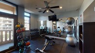 This fitness center is designed to support a comprehensive workout experience within a compact, well-organized layout. The space is divided into two interconnected zones separated by elegant French doors with frosted glass panels, allowing for a sense of openness while maintaining distinct workout areas.

The primary exercise area features durable rubber flooring throughout and is equipped with a wide range of free weights, including a full rack of dumbbells and an assortment of kettlebells and medicine balls organized on dedicated racks. An adjustable workout bench anchors the strength training zone, complemented by a large wall mirror that enhances the sense of space. Stability balls are neatly stacked along the mirrored wall, adding to the versatility of available equipment.

The cardio section houses a True treadmill, a recumbent bike, and an elliptical machine, positioned near large windows that invite natural light and offer views of the surrounding urban environment. A wall-mounted flat-screen television keeps users entertained during workouts, while a ceiling fan ensures comfortable airflow throughout.

A standout design feature is the bold motivational typography mural covering one full wall, displaying words such as "commitment," "focus," "believe," and "results" in a striking teal and gray palette. Recessed lighting and ceiling fans complete the practical, energizing atmosphere of this fitness space.