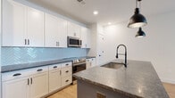 This single-floor residence showcases a thoughtfully designed interior with a cohesive modern aesthetic throughout. The centerpiece of the space is an open-concept kitchen that commands attention with its striking teal-blue island topped with dark granite countertops and an undermount sink fitted with a matte black pull-down faucet. White shaker-style cabinetry lines the walls from floor to ceiling, complemented by a light blue herringbone tile backsplash that adds a refined textural element. Stainless steel appliances — including a French door refrigerator, electric range, and built-in microwave — complete the kitchen's functional appeal. Two matte black pendant lights hang above the island, reinforcing the space's contemporary design language. Light-toned hardwood-style flooring flows throughout the main living areas, creating a warm, cohesive foundation. The bathroom continues the modern theme with white cabinetry, a dark granite vanity top, a bold black-framed mirror, matte black fixtures, and a tiled tub-shower combination. Bedrooms feature plush neutral carpeting, ceiling fans, and windows that allow natural light to fill each room. Recessed lighting throughout ensures the space remains bright and well-illuminated, resulting in a clean, move-in-ready environment.