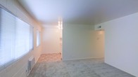 This single-floor space offers a clean, functional layout designed for comfortable everyday living. The main living area features neutral-toned carpet and bright white walls that lend a fresh, open atmosphere. Natural light filters in through a window fitted with blinds, illuminating the room and enhancing its airy feel. A wall-mounted HVAC unit provides climate control directly in the living space.

Transitioning into the adjacent area, the flooring shifts to durable tile, delineating the kitchen zone from the living space. The kitchen area is visible through an open pass-through layout, featuring white cabinetry and overhead lighting that keeps the space bright and practical. A central light fixture anchors this transitional zone.

The space also includes a visible HVAC vent near the ceiling and a wall thermostat, indicating a climate control system throughout. A doorway with overhead lighting leads further into the unit, suggesting additional rooms or storage beyond what is immediately visible.

Overall, the space presents a straightforward, well-maintained interior with neutral finishes, functional features, and a cohesive flow between the living and kitchen areas, making it a practical and comfortable single-level space.