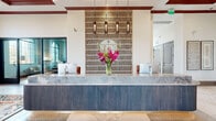 Located in Scottsdale, Arizona, this grand lobby hallway is a showcase of refined Southwestern architecture and upscale interior design. The single-floor space spans a generous footprint, offering an open, flowing layout that guides visitors through a series of elegantly connected areas. At the heart of the entrance is a striking reception desk finished in fluted blue cabinetry topped with a veined marble surface, set against a dramatic gold lattice screen wall accented by pendant lighting. Rich wood-trimmed coffered ceilings with a statement candle-style chandelier define the entry zone, while large arched windows flood the space with natural light and frame views of the Arizona landscape. Wide-format stone tile flooring in a chevron pattern runs throughout, complementing the warm wood floors in select areas. Graceful arched passageways connect hallway segments, each adorned with curated artwork, built-in shelving displaying artisan ceramics, and carefully placed seating vignettes anchored by patterned area rugs. Tall terracotta urns with tropical plants add organic warmth, while a sweeping iron-railed staircase with decorative tile risers serves as a dramatic architectural focal point. The overall ambiance is welcoming yet sophisticated, balancing Mediterranean-inspired architecture with culturally rich, desert-contemporary furnishings.