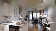 This single-story residence showcases an open-concept layout that seamlessly connects the kitchen, dining, and living areas into one cohesive, functional space. The kitchen is outfitted with granite countertops, a stainless steel dishwasher, and a breakfast bar that doubles as a casual dining surface, making it ideal for both cooking and entertaining. Track lighting overhead provides adjustable illumination throughout the space.

The living area is generously proportioned, featuring rich hardwood-style flooring that runs throughout the main living zone, anchored by a large area rug. A ceiling fan adds both style and practicality. Large windows dressed with drapes and blinds allow an abundance of natural light to fill the room, and a glass door opens directly to a private balcony, extending the living space outdoors.

The neutral warm-toned walls provide a versatile backdrop that complements a range of interior styles. A glimpse into an adjacent bedroom reveals continued consistency in finishes and design. Overall, the space presents a well-balanced combination of modern aesthetics and practical functionality.