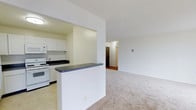 This single-floor residence offers a clean and functional layout designed for comfortable everyday living. The open-concept floor plan connects the kitchen and living area in a seamless flow, creating a sense of spaciousness throughout. The kitchen is equipped with white cabinetry, white appliances including a range and microwave, and dark countertops that provide a subtle contrast to the bright, neutral surroundings. A half-wall peninsula serves as a natural divider between the kitchen and the living space, adding definition without closing off the room. The living area features soft, neutral wall-to-wall carpeting that contributes to a warm and welcoming ambiance. Smooth white walls and consistent overhead lighting keep the interior feeling bright and airy. A hallway visible from the main living space leads further into the unit, hinting at additional private rooms beyond. The overall design emphasizes practicality and simplicity, with a cohesive neutral color palette of whites and beiges that offers a versatile backdrop for a variety of interior styles. The space is well-suited for those seeking a straightforward, move-in ready layout with an efficient use of its footprint.
