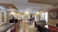 This single-story amenity space is thoughtfully designed to serve as a communal gathering hub. The interior is finished with warm beige walls, hardwood-style flooring, and a combination of tiled and carpeted areas that delineate functional zones throughout. Tray ceilings with recessed and track lighting add architectural depth and provide well-distributed illumination across the space.

The lounge area is furnished with a mix of patterned fabric sofas, leather seating, and accent chairs arranged around low coffee tables, creating a relaxed and welcoming atmosphere. Multiple sets of glass French doors line one wall, flooding the interior with natural light and offering direct views and access to an outdoor pool and patio area, where lounge chairs and seating are visible.

A café-style seating section features round metallic bistro tables paired with high-back bar stools, positioned near a coffee and beverage station. This area connects seamlessly to a full kitchen equipped with dark granite countertops, warm wood cabinetry, a decorative tile backsplash, stainless steel appliances including a full-size refrigerator, and recessed tray ceiling lighting. A service window connects the kitchen to the adjacent lounge, enhancing the space's functionality for community events and gatherings.