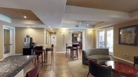 This single-story amenity space is thoughtfully designed to serve as a communal gathering hub. The interior is finished with warm beige walls, hardwood-style flooring, and a combination of tiled and carpeted areas that delineate functional zones throughout. Tray ceilings with recessed and track lighting add architectural depth and provide well-distributed illumination across the space.

The lounge area is furnished with a mix of patterned fabric sofas, leather seating, and accent chairs arranged around low coffee tables, creating a relaxed and welcoming atmosphere. Multiple sets of glass French doors line one wall, flooding the interior with natural light and offering direct views and access to an outdoor pool and patio area, where lounge chairs and seating are visible.

A café-style seating section features round metallic bistro tables paired with high-back bar stools, positioned near a coffee and beverage station. This area connects seamlessly to a full kitchen equipped with dark granite countertops, warm wood cabinetry, a decorative tile backsplash, stainless steel appliances including a full-size refrigerator, and recessed tray ceiling lighting. A service window connects the kitchen to the adjacent lounge, enhancing the space's functionality for community events and gatherings.
