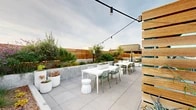 This expansive rooftop terrace offers a sophisticated outdoor retreat designed with both aesthetics and functionality in mind. The space is organized into several distinct zones, each serving a unique purpose while contributing to a cohesive, modern design language. Large-format porcelain pavers provide a clean, continuous ground surface throughout, lending a polished, contemporary feel underfoot.

A covered lounge area features a warm plywood ceiling, steel-frame columns, and floor-to-ceiling glass panels that frame panoramic views of the surrounding cityscape and rolling hills. String lights overhead add an inviting ambiance that transitions seamlessly from day to evening. An open-air dining zone offers white tables with sage-green seating, surrounded by large white planters filled with sculptural plants and ornamental grasses.

A steel pergola anchors a second lounge area furnished with deep-seated sectional sofas, ideal for relaxed gatherings. Raised concrete planters host a curated mix of olive trees, succulents, flowering perennials, and trailing greenery, creating natural privacy screens and visual texture. A living green roof runs along the perimeter railing, layered with low-growing sedum and gravel edging.

Panoramic views extend across dense urban neighborhoods to prominent hilltop landmarks, visible from nearly every vantage point on the terrace.