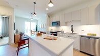 This contemporary single-floor residence offers a thoughtfully designed open-concept layout that seamlessly connects the kitchen, dining, and living areas. The kitchen is a standout feature, showcasing flat-front white cabinetry, stainless steel appliances including a refrigerator, dishwasher, microwave, and range, complemented by a sleek white quartz countertop island with bar seating. Stylish pendant lighting adds a modern touch overhead, while light wood flooring runs throughout, creating a warm and cohesive aesthetic.

The living room is bright and welcoming, filled with natural light streaming through large sliding glass doors that open onto a private balcony, offering open outdoor views. The space is accented by a ceiling fan and warm, earthy tones.

The residence includes two well-appointed bedrooms, each featuring large windows that invite ample natural light, ceiling fans, and neutral carpeting. Both rooms offer a calm and restful ambiance.

The full bathroom is clean and functional, finished with large-format white tile flooring, a single vanity with a framed mirror, matte black fixtures, and a tub-shower combination. The overall design of this residence balances modern minimalism with warm, inviting elements throughout.