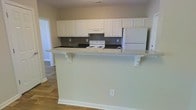 This two-story space offers a practical and well-organized layout designed to accommodate everyday living. The kitchen serves as a focal point, featuring white raised-panel cabinetry that extends to the upper walls, providing generous storage. Dark countertops contrast with the white cabinetry, while a gray tile backsplash adds a subtle textural detail. A white electric range and a full-size refrigerator complete the appliance package. A peninsula with a granite-style countertop extends outward, supported by decorative brackets, creating a breakfast bar ideal for casual dining or additional workspace. The warm-toned hardwood flooring runs throughout the main level, contributing to a cohesive and inviting atmosphere. Neutral wall tones throughout keep the space feeling open and versatile, offering a clean backdrop suitable for a variety of interior styles. The two-story layout suggests additional living or sleeping quarters on the upper level, expanding the overall functionality of the home. Overall, the space presents a straightforward, move-in-ready environment with classic design elements and practical features throughout.
