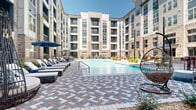 This resort-style pool area in Birmingham, Alabama, is a thoughtfully designed outdoor amenity space set within a curved, multi-story building that creates a semi-enclosed courtyard atmosphere. The centerpiece is a generously sized swimming pool with crystal-clear blue water, framed by an eye-catching herringbone-patterned paver deck in a blend of gray, red, and tan tones that adds visual interest and texture throughout the space.

The surrounding deck is well-appointed with cushioned lounge chairs, blue side tables, and market umbrellas, providing ample space for sunbathing and relaxation. Unique hanging egg chairs add a stylish, resort-inspired touch to seating areas nestled against the building's exterior. Colorful oversized ceramic planters and carefully maintained landscaping beds featuring shrubs, ornamental grasses, and flowering plants introduce vibrant greenery and natural elements throughout the courtyard.

The building enclosure features a contemporary mix of brick masonry, light panel siding, and dark-framed windows with private balconies above, creating a visually dynamic backdrop. Concrete walkways wind through the landscaped perimeter, connecting entry points and providing easy circulation. The open-sky design allows abundant natural light to fill the space, enhancing the bright, resort-like ambiance of this outdoor retreat.