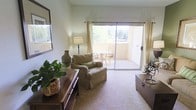 This single-floor residence features a welcoming living room designed for both comfort and functionality. The space is finished with wall-to-wall carpeting in a warm neutral tone, contributing to a cozy and inviting atmosphere throughout. A key highlight of the living room is the large sliding glass door flanked by sheer, gold-toned curtains, which floods the interior with abundant natural light and provides direct access to a private balcony. The balcony extends the living area outdoors and is furnished with a table and chairs, making it ideal for relaxed outdoor dining or casual seating while enjoying the surrounding greenery and tree views. Inside, the living room is tastefully arranged with a cushioned armchair accented by a decorative pillow, a classic wood credenza adorned with a potted plant and decorative accessories, and a framed wall mirror that enhances the sense of space. A traditional floor lamp adds warm ambient lighting to complement the natural light. The overall design aesthetic is understated and comfortable, with neutral walls and warm wood tones creating a harmonious and livable environment suited to everyday use.