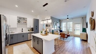 This well-appointed single-floor residence offers a thoughtfully designed open-concept layout where the kitchen flows seamlessly into the living area, creating an inviting and connected living experience. The kitchen is a standout feature, boasting two-tone cabinetry with white upper cabinets and slate-gray lower cabinets, white quartz countertops, a classic subway tile backsplash, stainless steel appliances, and a central island with bar seating — ideal for both cooking and casual dining. Pendant lighting and recessed fixtures brighten the space throughout. The living room offers a relaxed ambiance with warm hardwood-style flooring, a ceiling fan, large windows, and a private exterior door that invites natural light and outdoor views. Two bedrooms provide comfortable retreats; one features a bold teal accent wall and natural light from large windows, while the second is finished in warm neutral tones. Both bathrooms are finished with gray vanity cabinetry and white quartz countertops. The full bath includes a soaking tub and shower combination with large-format tile surround, while the second bath features a clean, minimalist design. Consistent hardwood-style flooring and a neutral color palette tie the entire residence together in a cohesive, modern aesthetic.