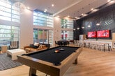 Located at 500 Race St in San Jose, CA, this single-floor amenity space at Mosaic Apartments offers a generously sized, open-concept layout designed to serve as a dynamic communal hub. The space is immediately defined by its dramatic double-height ceilings and expansive floor-to-ceiling glass walls that flood the interior with natural light and provide views of the surrounding outdoor courtyard and landscaping. Light-toned hardwood flooring runs throughout, lending warmth and visual continuity across the entire space.

The main entrance features a grand set of glass double doors framed by towering glazed panels, creating an airy, welcoming arrival experience. A branded Mosaic welcome mat marks the threshold. The open layout is thoughtfully divided into distinct functional zones: a lounge area with contemporary seating including a leather sofa and armchairs, a billiards area anchored by a full-size pool table beneath dramatic spherical pendant lights, and a sleek linear fireplace with a wall-mounted TV set against deep charcoal tile.

A co-working and dining zone includes long communal tables with white café chairs, pendant globe lighting, and a bold graphic accent wall with dual mounted screens. A kitchenette with white cabinetry and appliances supports this area. Additional seating nooks with colorful accent chairs complete the space, making it a versatile and stylish environment for work, recreation, and socializing.
