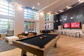 Located at 500 Race St in San Jose, CA, this single-floor amenity space at Mosaic Apartments offers a generously sized, open-concept layout designed to serve as a dynamic communal hub. The space is immediately defined by its dramatic double-height ceilings and expansive floor-to-ceiling glass walls that flood the interior with natural light and provide views of the surrounding outdoor courtyard and landscaping. Light-toned hardwood flooring runs throughout, lending warmth and visual continuity across the entire space.

The main entrance features a grand set of glass double doors framed by towering glazed panels, creating an airy, welcoming arrival experience. A branded Mosaic welcome mat marks the threshold. The open layout is thoughtfully divided into distinct functional zones: a lounge area with contemporary seating including a leather sofa and armchairs, a billiards area anchored by a full-size pool table beneath dramatic spherical pendant lights, and a sleek linear fireplace with a wall-mounted TV set against deep charcoal tile.

A co-working and dining zone includes long communal tables with white café chairs, pendant globe lighting, and a bold graphic accent wall with dual mounted screens. A kitchenette with white cabinetry and appliances supports this area. Additional seating nooks with colorful accent chairs complete the space, making it a versatile and stylish environment for work, recreation, and socializing.