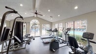 This single-story fitness center in Houston, TX (77034) offers a thoughtfully designed workout space filled with both cardio and strength training equipment. The facility features vaulted ceilings with exposed beams and a textured finish, fitted with recessed lighting and ceiling fans to maintain a comfortable environment during workouts. Large windows, including a prominent arched window, line the walls and flood the space with natural light while providing views of the surrounding outdoor pool area and landscaping. The cardio section is well-stocked with Life Fitness treadmills, elliptical trainers, and stationary and recumbent bikes, all positioned to take advantage of the outdoor views. A dedicated strength training area includes a multi-station cable machine, adjustable weight benches, and free weights. Full-length mirrors are mounted along one wall, visually expanding the space and allowing users to monitor their form. The durable rubber flooring provides cushioning and traction throughout. French doors on one end of the facility provide direct access to the exterior. The bright, open layout and abundance of equipment make this fitness center a comprehensive and functional space for a full-body workout routine.