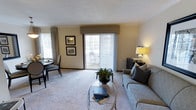 This single-floor residence offers a welcoming and thoughtfully arranged living area finished in warm neutral tones throughout. The main living space is anchored by plush wall-to-wall carpeting and features a large sliding glass door fitted with vertical blinds and a decorative valance, allowing generous natural light to fill the room while providing direct access to a private outdoor balcony. The balcony, visible through the glass door, offers a pleasant outdoor retreat. A comfortable seating area centers around a circular glass-top coffee table, complemented by a classic sofa with decorative pillows. A credenza with table lamp adds a warm, ambient glow to the space. Adjacent to the main seating area, a compact dining space accommodates a small round table and chairs, positioned near a second window dressed with patterned curtains, further enhancing the natural brightness of the room. Framed artwork adorns the walls, contributing to the polished, move-in-ready feel of the space. The overall ambiance is calm and inviting, with consistent warm tones and tasteful furnishings that create a comfortable and functional single-level living environment.
