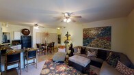 This single-story space showcases an open-concept layout that seamlessly connects the living and dining areas, creating a cohesive and functional environment. The living area is anchored by a large, plush sectional sofa in a neutral gray tone, paired with a tufted ottoman and accented with decorative throw pillows. A bold, large-scale mixed-media artwork serves as a striking focal point on the main wall, adding color and visual interest to the space. A patterned area rug with warm floral motifs defines the seating zone and adds texture underfoot.

The dining area, situated adjacent to the living room, features a dark wood dining table surrounded by matching chairs, with a decorative floral arrangement as a centerpiece. Ceiling fans in both areas provide practical air circulation while contributing to the overall design. A chaise lounge and decorative floor lamp bridge the two zones, enhancing the transitional flow. The light-toned tile flooring runs throughout, complementing the warm, cream-colored walls. The overall ambiance is inviting and well-appointed, with thoughtful décor choices that balance comfort and style.