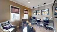 This single-floor space combines a functional business center with a comfortable lounge area, creating a versatile environment suited for both work and relaxation. The room is finished with warm taupe-toned walls accented by crisp white crown molding, lending a polished, professional aesthetic throughout.

Natural light fills the space through two windows dressed with rich, textured roman shades in deep plum tones, adding a layer of warmth and sophistication. A dedicated workstation runs along one wall, featuring a dark granite-topped desk equipped with desktop computers and office chairs, making it an efficient workspace for multiple users simultaneously. Above the workstation, a series of contemporary pendant lights with matte black fixtures provides focused task lighting, complemented by a row of modern framed artwork.

The lounge area offers comfortable upholstered armchairs in a neutral grey tone, arranged around a sleek round coffee table adorned with an elegant floral arrangement. Soft, patterned carpeting underfoot ties the two zones together cohesively, contributing to the room's overall refined and welcoming ambiance.