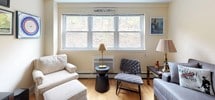 This single-floor residence offers a thoughtfully arranged layout with distinct living, sleeping, dining, and kitchen areas. The living room is anchored by a plush gray sectional sofa beneath a striking large-format black-and-white New York City cityscape canvas, complemented by warm hardwood flooring and a decorative sunburst wall accent. Natural light filters in through double-hung windows, creating a bright and inviting atmosphere. An adjoining seating nook features a comfortable lounge chair and loveseat arranged around a sleek pedestal side table, with a large window framing a verdant wooded view and a Morristown map print adding local character. The bedroom is generously sized, accommodating a king-sized bed with a tufted leather headboard, flanked by a classic wooden side table. A wall-mounted flat-screen television, open clothing rack, and a tall bookshelf provide functional storage and entertainment options. The kitchen is well-equipped with warm wood cabinetry, granite countertops, recessed lighting, tile flooring, and a black refrigerator. A butcher-block breakfast bar with metal bar stools offers a casual dining spot. The bathroom features neutral tile throughout, a white vanity with a granite countertop, and a shower-tub combination with glass sliding doors, completing this efficiently designed and personalized residence.