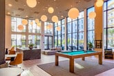 Located at 1350 S Bascom Avenue in San Jose, California, this two-floor amenity clubhouse is a thoughtfully designed social and recreational hub. The main floor commands immediate attention with its soaring double-height ceilings and an expansive wall of floor-to-ceiling windows that frame sweeping views of a resort-style pool courtyard lined with palm trees and lounge chairs. Oversized globe pendant lights cascade from above, illuminating light wood-look flooring and a central billiards table that anchors the room. A built-in bar counter with vibrant yellow stools, teal banquette seating, and a large-screen TV create a lively entertainment zone, while a lounge area with plush sofas invites relaxed conversation. A striking open-tread staircase with steel railings and a decorative rock feature at its base serves as both a functional and sculptural centerpiece. Curated wall art, including bold fan-shaped textile pieces, adds artistic character throughout. The second floor offers a more intimate game and lounge area, complete with a ping-pong table, hexagonal tile flooring, a kitchenette with dark marble counters, teal booth seating, and casual dining arrangements. Together, both levels deliver a well-rounded, modern amenity experience.