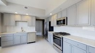 Located at 50 W Broad St in the heart of downtown Columbus, Ohio, this single-floor space offers a thoughtfully designed interior with contemporary finishes throughout. The kitchen serves as a central highlight, showcasing gray shaker-style cabinetry that extends to the ceiling, providing generous storage capacity. Crisp white countertops contrast elegantly with the cabinetry, creating a clean, modern aesthetic. Stainless steel appliances, including a gas range, built-in microwave, French door refrigerator, and dishwasher, outfit the kitchen with full functionality. A gold-toned faucet at the sink adds a refined accent detail. Recessed lighting illuminates the space evenly, complementing the light-toned tile flooring that runs throughout. The layout offers an efficient, galley-style flow, with ample counter space on both sides for meal preparation. A doorway visible from the kitchen suggests a well-connected floor plan, allowing for easy movement throughout the space. The overall design balances practicality with modern style, making it well-suited for everyday living in a prime urban location.