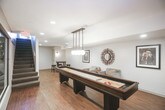 This thoughtfully designed lower-level space offers a range of functional and recreational areas unified by warm hardwood-style flooring, neutral wall tones, and recessed lighting throughout. The lounge area provides a comfortable setting with upholstered seating, a decorative sunburst mirror, and framed artwork adorning the clean white walls, creating a relaxed and inviting atmosphere.

Adjacent to the lounge, a dedicated dining nook features a round bar-height table paired with dark stools, accented by a wall sconce and additional framed art. The space is well-suited for casual gatherings or entertaining.

The game room is a standout feature, anchored by a full-length shuffleboard table beneath globe pendant lighting. Large glass windows and walls visually connect this area to the surrounding rooms, maintaining an open and airy feel despite the lower-level setting.

Enclosed behind floor-to-ceiling glass panels, the fitness center is equipped with cardio machines and exercise equipment, offering both privacy and visibility. A staircase leads back to the upper level, completing this well-appointed and versatile lower-level layout.