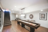 This thoughtfully designed lower-level space offers a range of functional and recreational areas unified by warm hardwood-style flooring, neutral wall tones, and recessed lighting throughout. The lounge area provides a comfortable setting with upholstered seating, a decorative sunburst mirror, and framed artwork adorning the clean white walls, creating a relaxed and inviting atmosphere.

Adjacent to the lounge, a dedicated dining nook features a round bar-height table paired with dark stools, accented by a wall sconce and additional framed art. The space is well-suited for casual gatherings or entertaining.

The game room is a standout feature, anchored by a full-length shuffleboard table beneath globe pendant lighting. Large glass windows and walls visually connect this area to the surrounding rooms, maintaining an open and airy feel despite the lower-level setting.

Enclosed behind floor-to-ceiling glass panels, the fitness center is equipped with cardio machines and exercise equipment, offering both privacy and visibility. A staircase leads back to the upper level, completing this well-appointed and versatile lower-level layout.